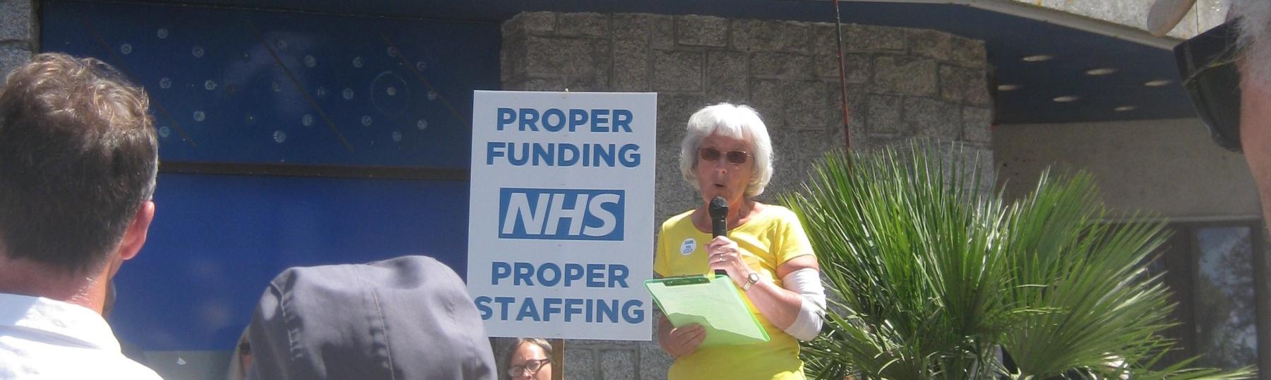 Woman speaking to a crowd outdoors. Placard reads "Proper funding. NHS. Proper staffing.