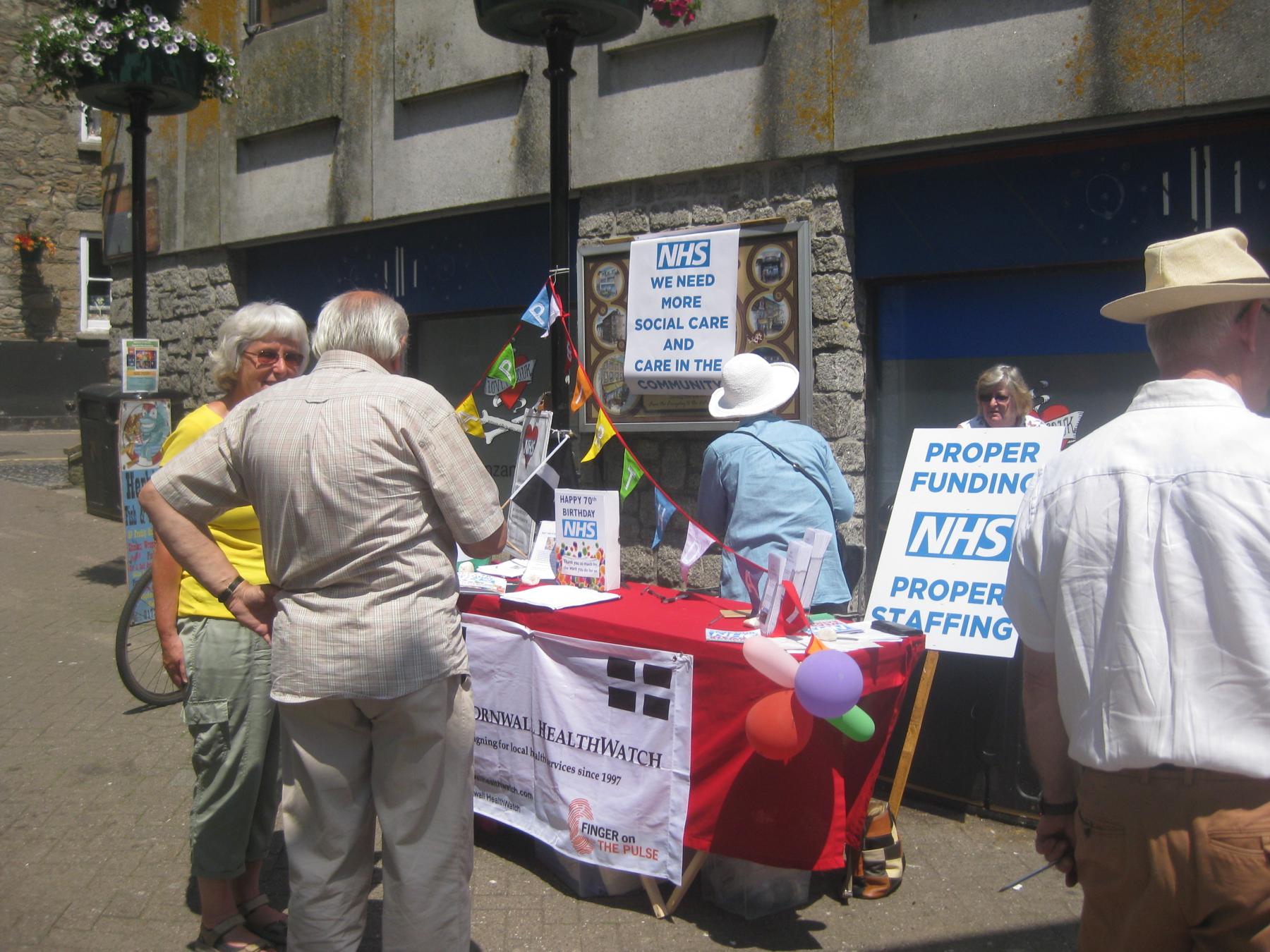 People round West Cornwall Healthwatch stall on Causewayhead. Placards read "Proper Funding. NHS. Proper Staffing" and "We need more social care and care in the community"